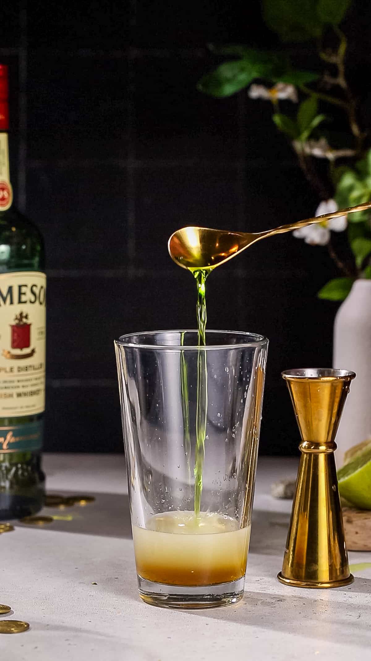 Hand using a bar spoon to add Midori liqueur to a cocktail shaker.