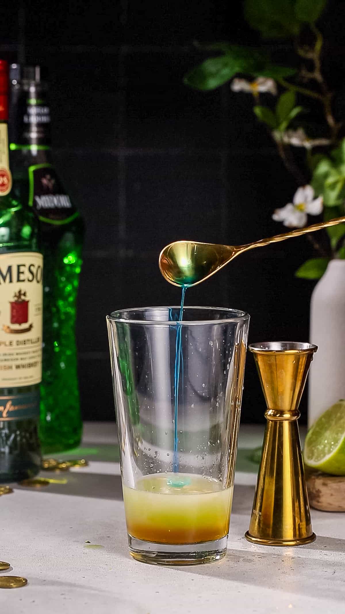 Hand using a bar spoon to add Blue Curacao liqueur to a cocktail shaker.