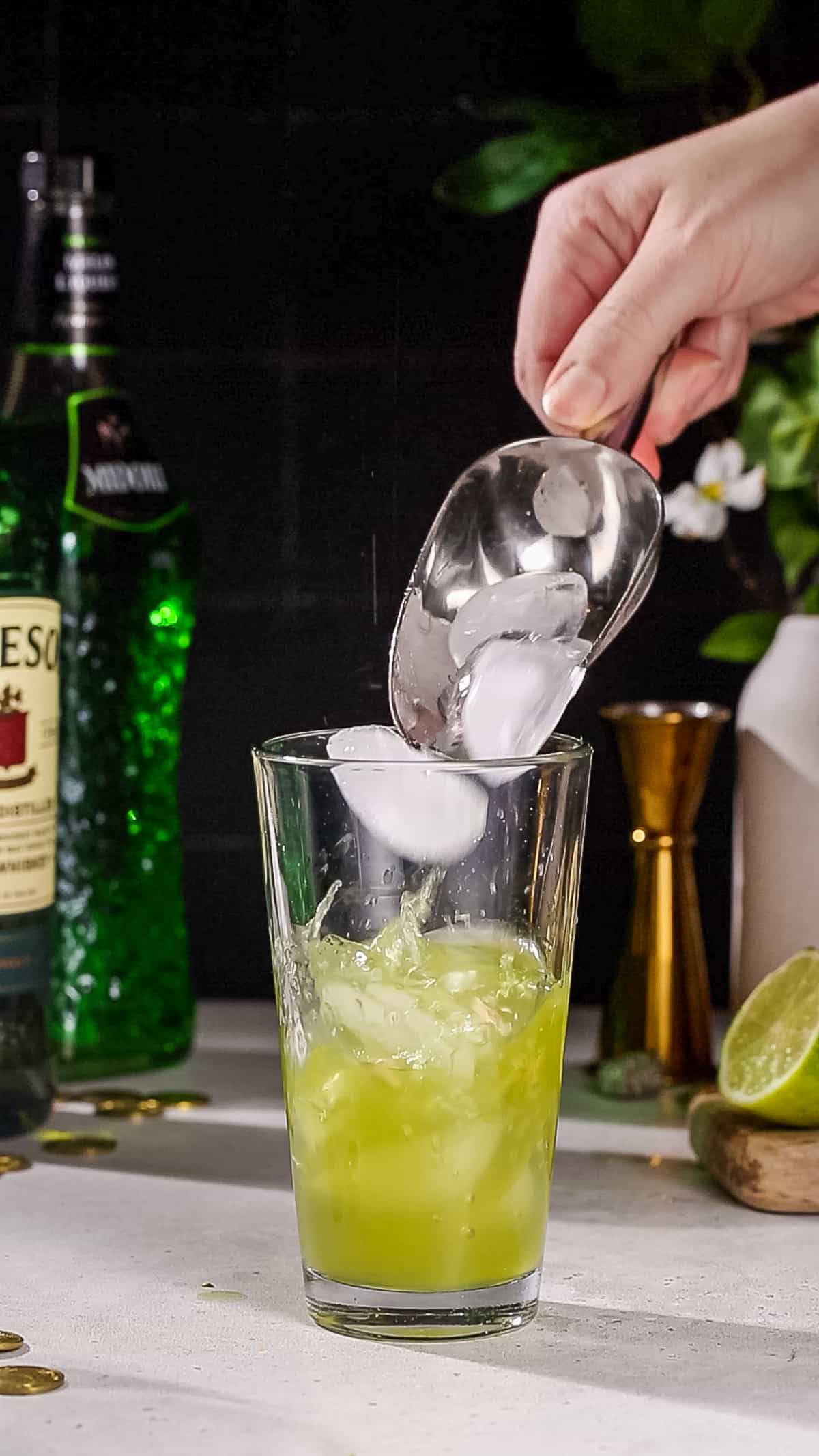 Hand adding ice to a cocktail shaker filled with green liquid.