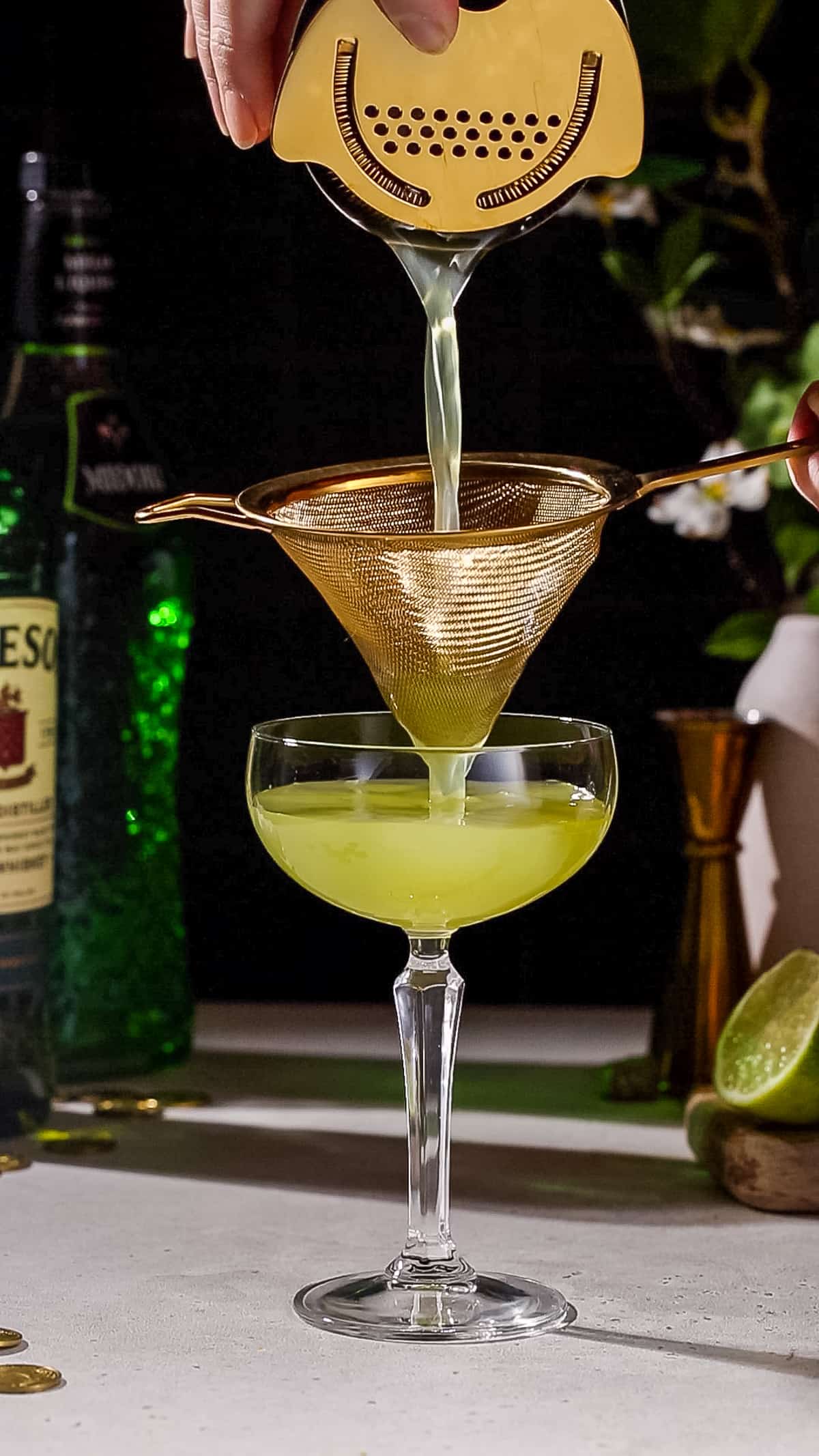 Hand straining a green colored cocktail into a coupe glass.