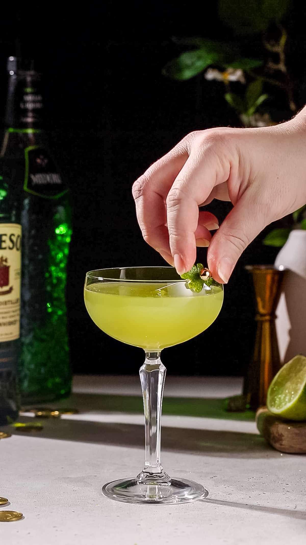 Hand adding a lime peel in the shape of a shamrock to a green colored cocktail as a garnish.