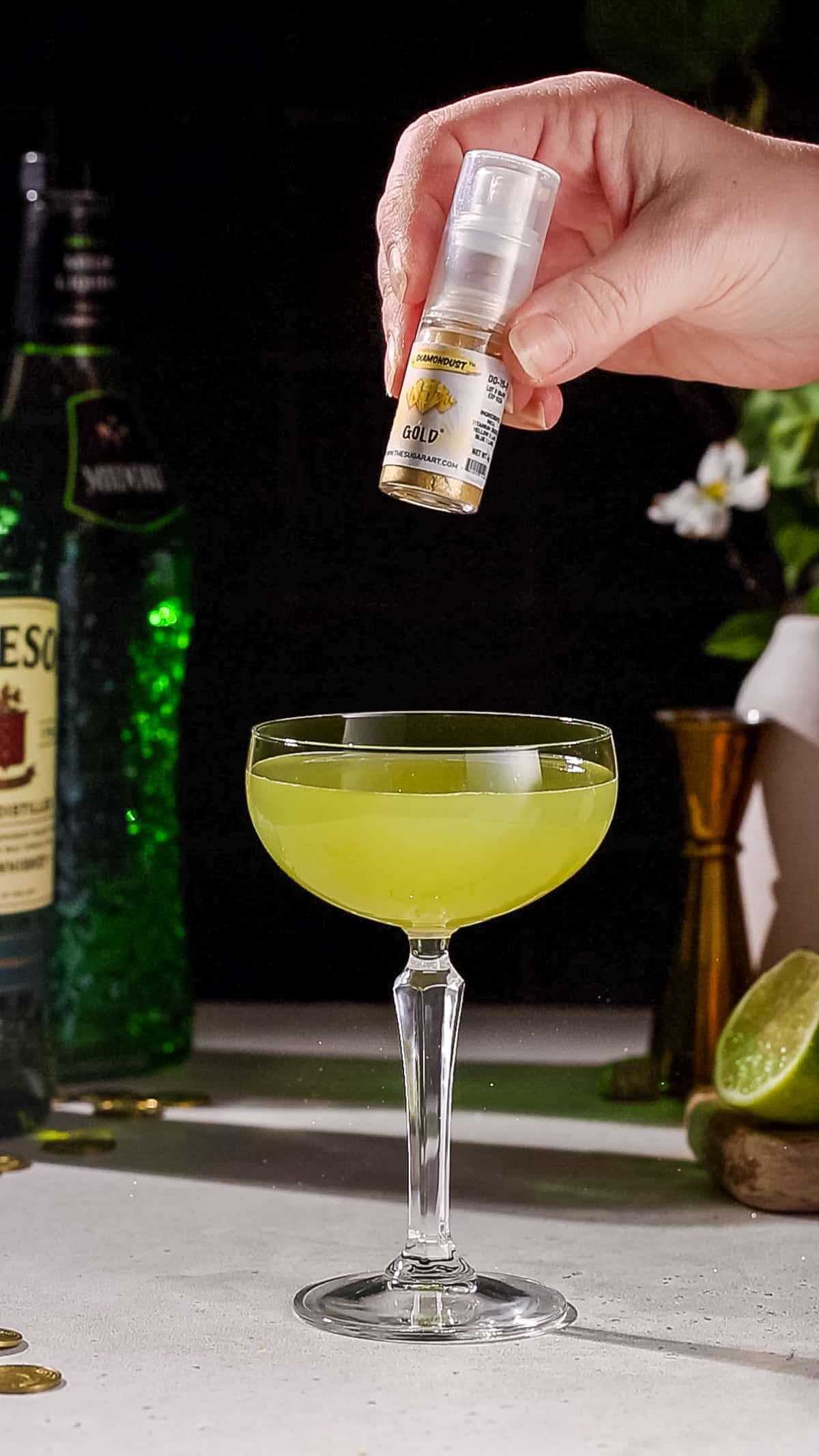 Hand showing a bottle of gold edible glitter above a green cocktail.