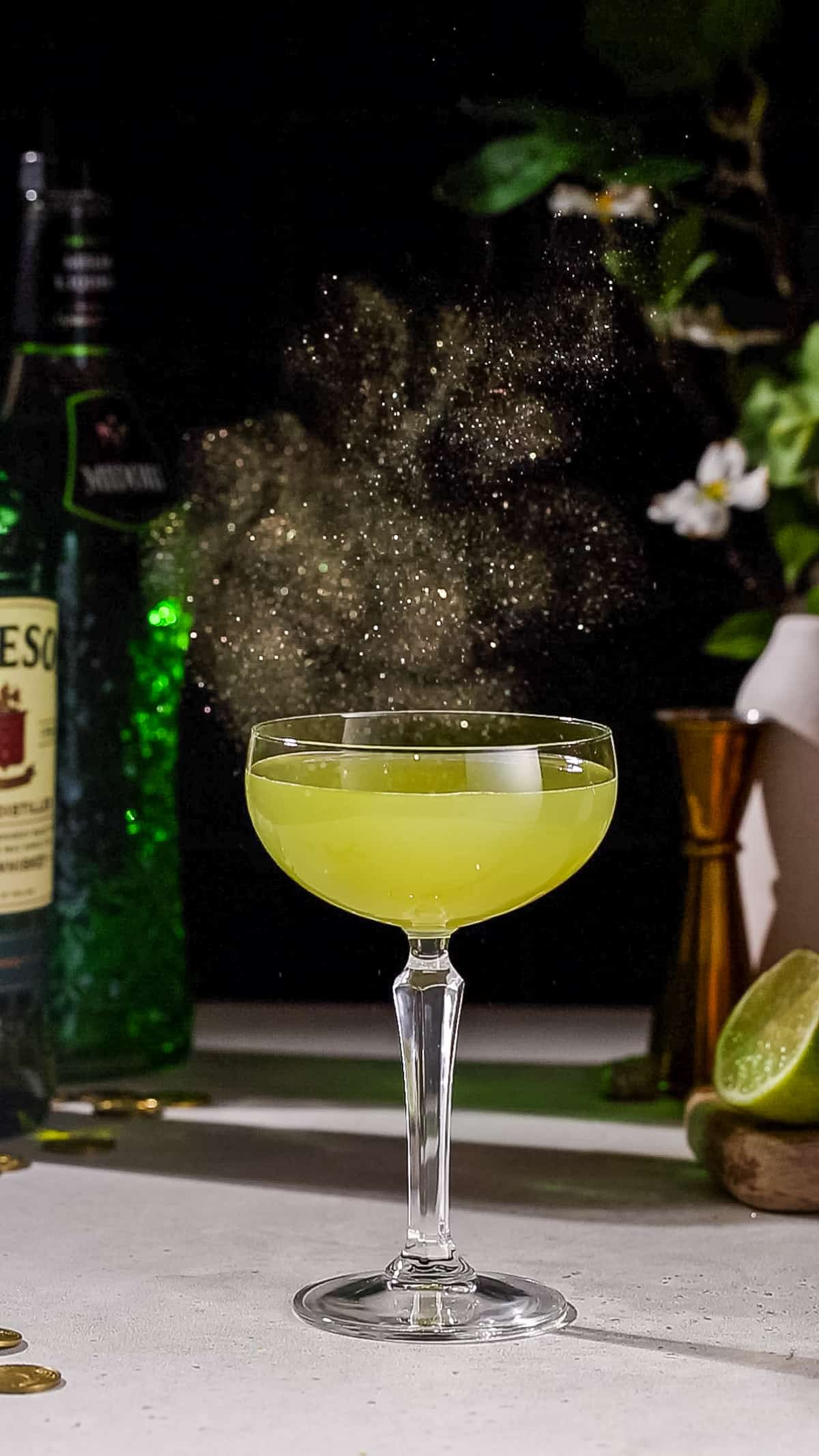 A cloud of gold glitter hangs in the air above a green colored cocktail in a coupe glass.