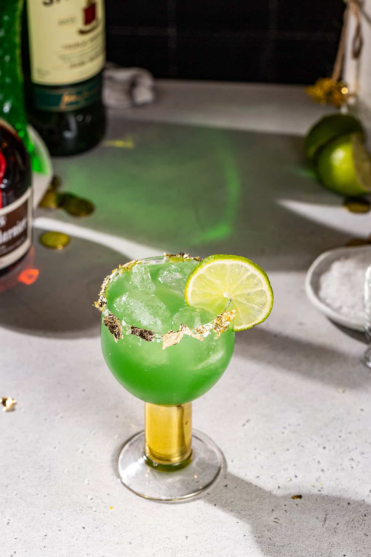 Overhead view of a green colored Irish Margarita cocktail. In the background is a dish of salt, a cut lime, and some bottles of alcohol used to make the drink.