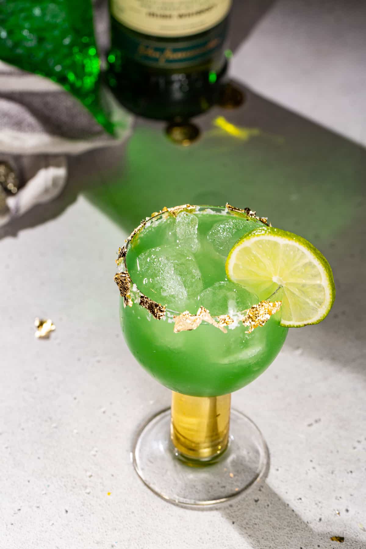 Overhead view of a green colored Irish Margarita cocktail with a lime and edible gold garnish. Some liquor bottles are in the background.