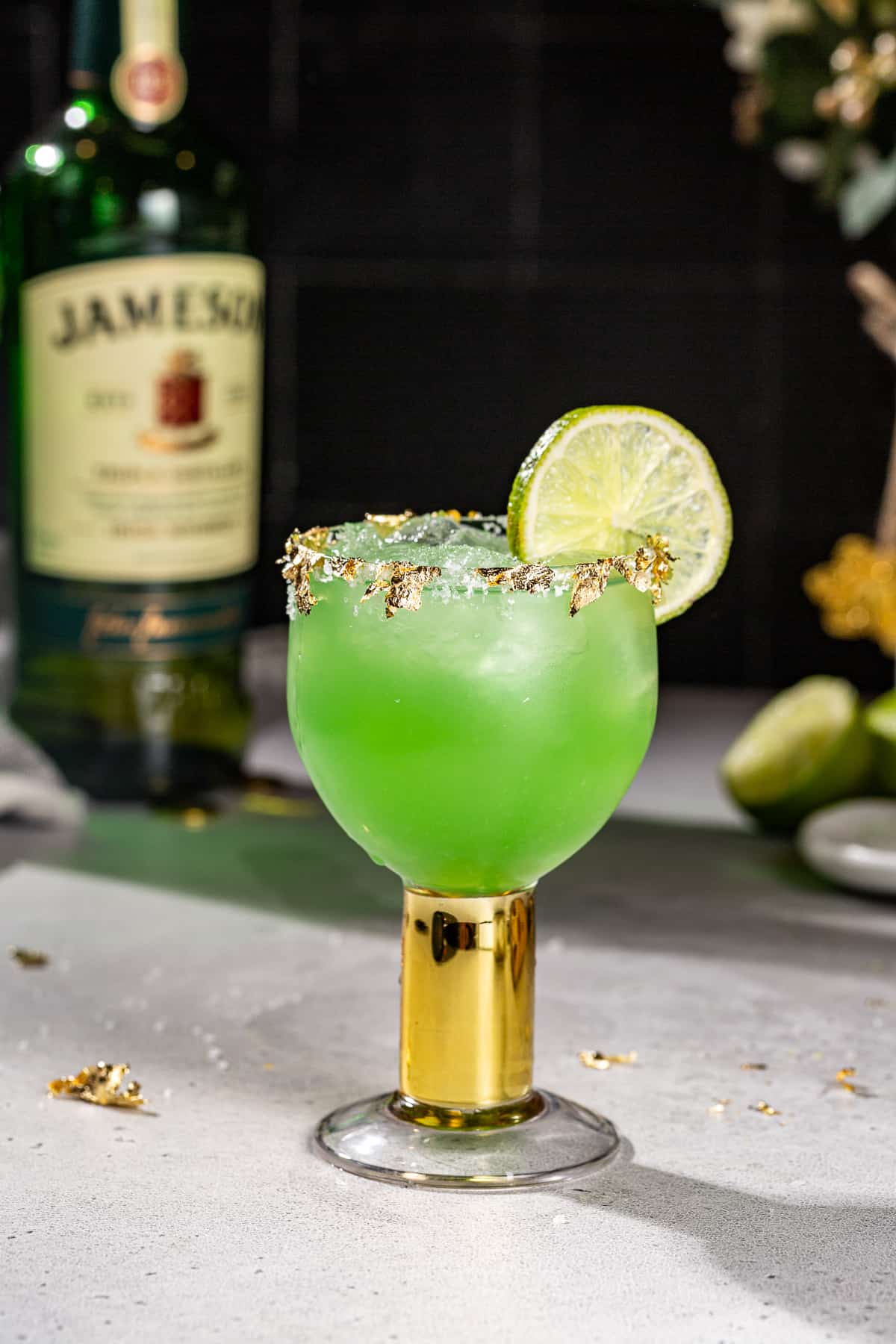 Side view of a green colored Irish Margarita cocktail with edible gold leaf, salt rim and a lime wheel garnish. A bottle of Jameson Irish whiskey is in the background.