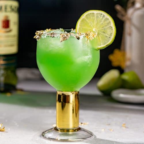 Side view of a green colored Irish Margarita. The drink is garnished with gold leaf, salt and a lime.