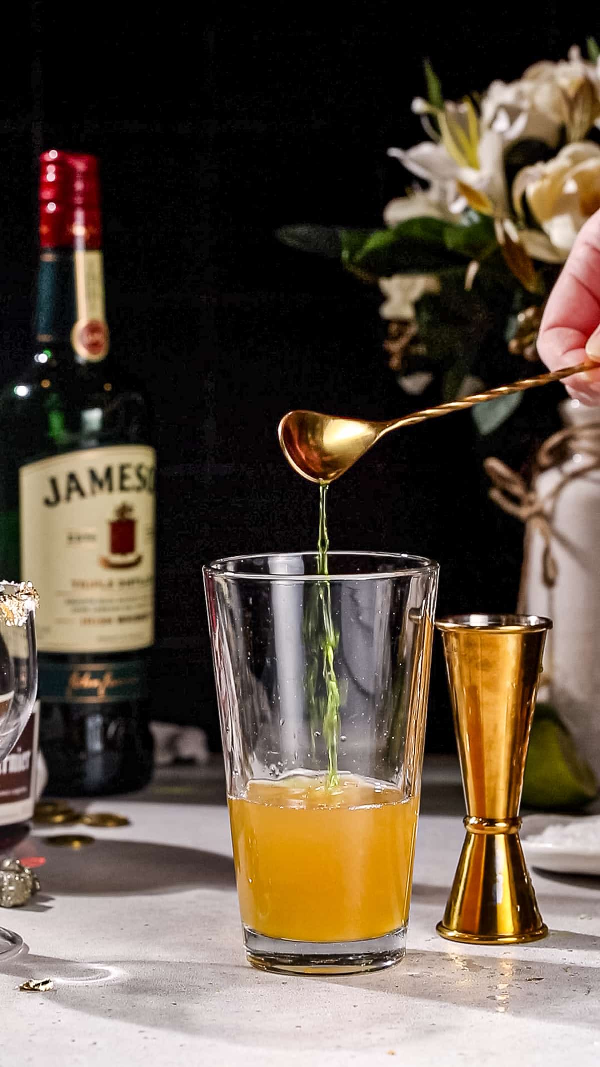 Hand using a bar spoon to add Midori liqueur to a cocktail shaker filled with orange liquid.