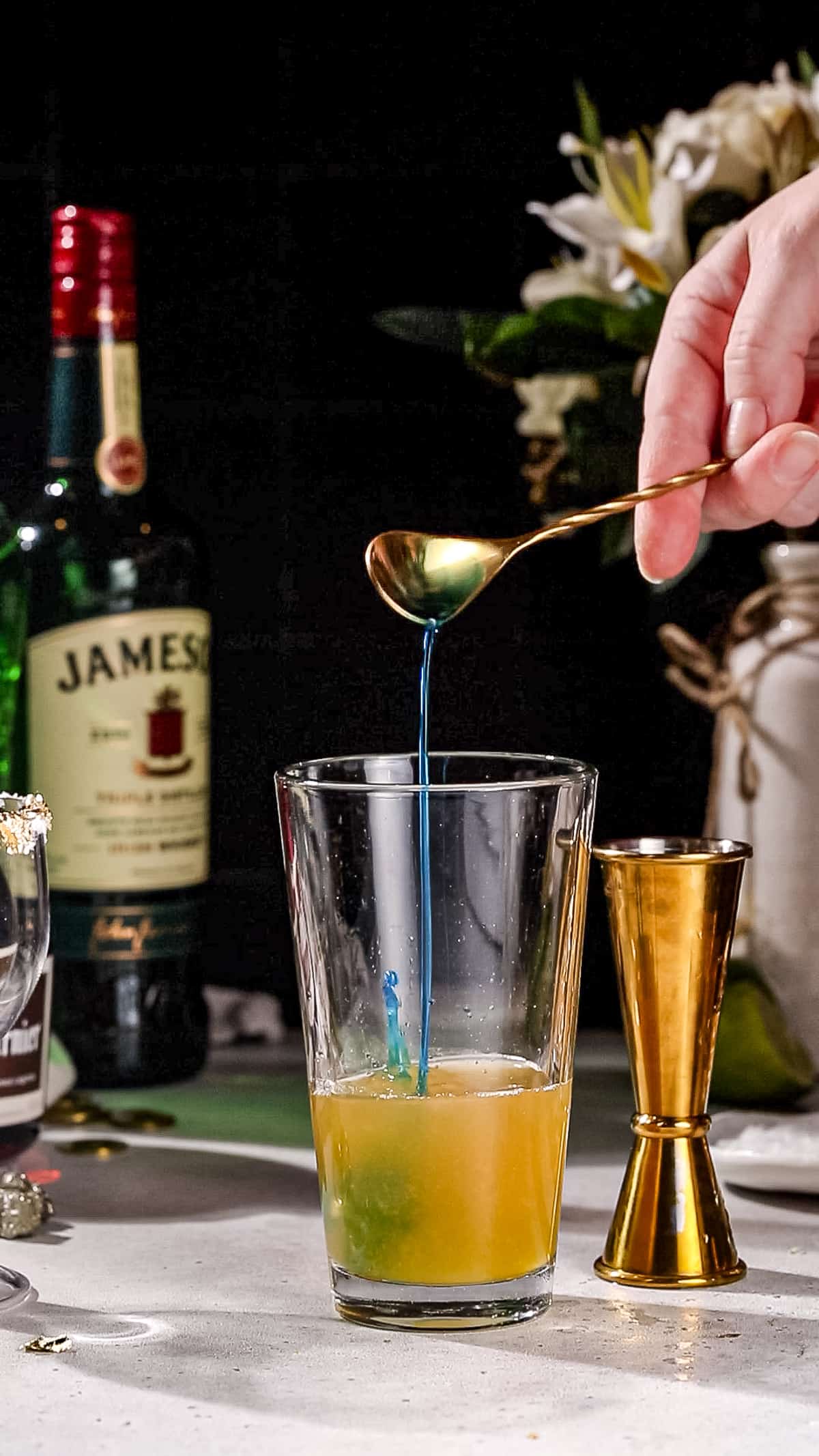 Hand using a bar spoon to add Blue Curacao liqueur to a cocktail shaker filled with orange liquid.