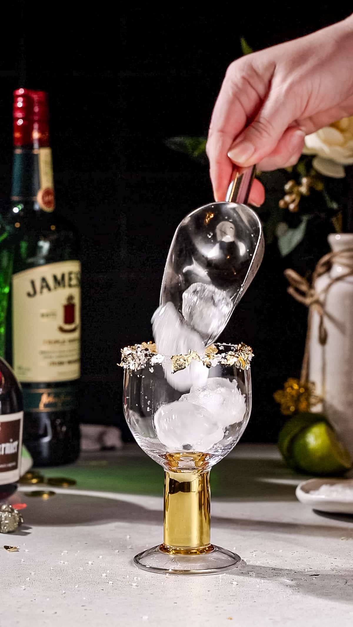 Hand using an ice scoop to add ice cubes to a gold-stemmed cocktail glass that is rimmed with salt and gold leaf.