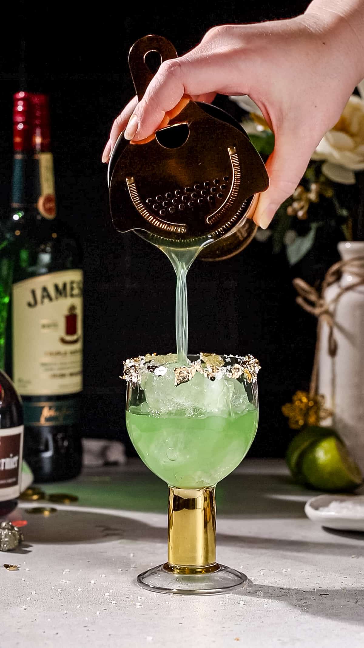 Hand straining a green colored drink into a cocktail glass.