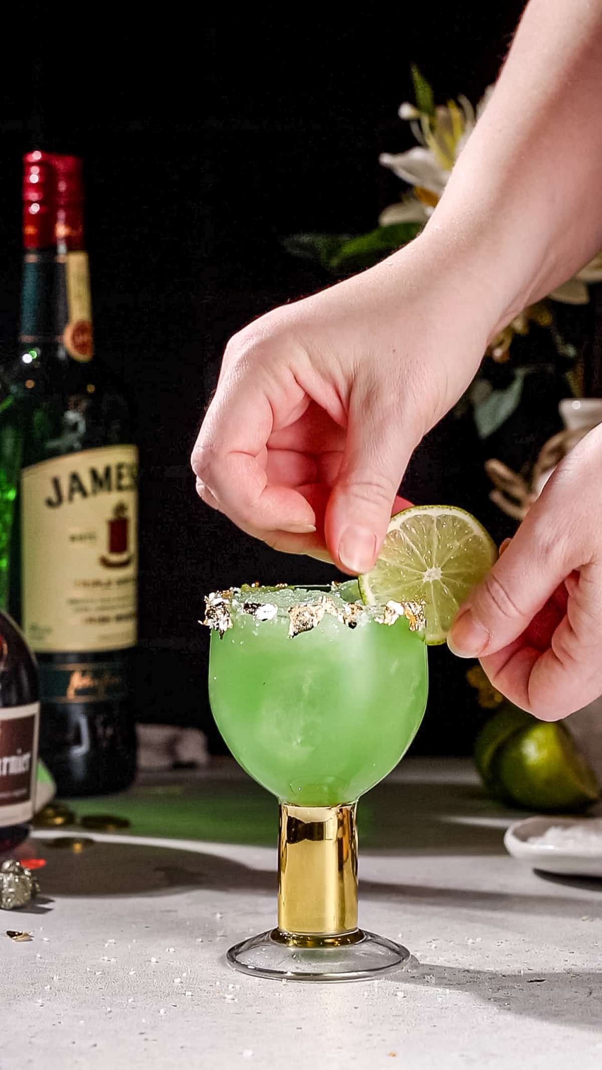 Hand adding a lime slice to a cocktail glass filled with green liquid.