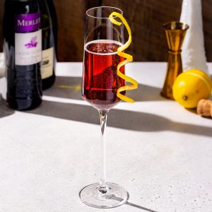 Kir Royale cocktail in a champagne flute with lemon peel garnish. A lemon along with a gold jigger and a champagne cork are in the background, along with a bottle of champagne and creme de cassis.