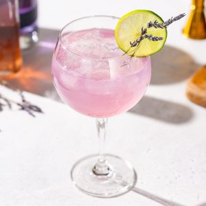 Slightly overhead photo of a Lavender Lime gin spritz cocktail. The drink is a pinkish purple color and garnished with a lime slice and lavender sprigs.