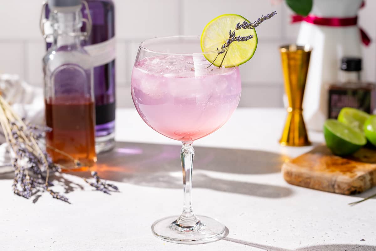 Lavender Lime gin spritz cocktail on a countertop. The drink is a purple color and garnished with a lime slice and lavender sprigs. In the background are ingredients to make the drink, including a bottle of Empress gin, a bottle of lavender syrup, a fresh lime, and a bottle of lavender bitters. There is also a cutting board, a vase of flowers and a gold jigger.