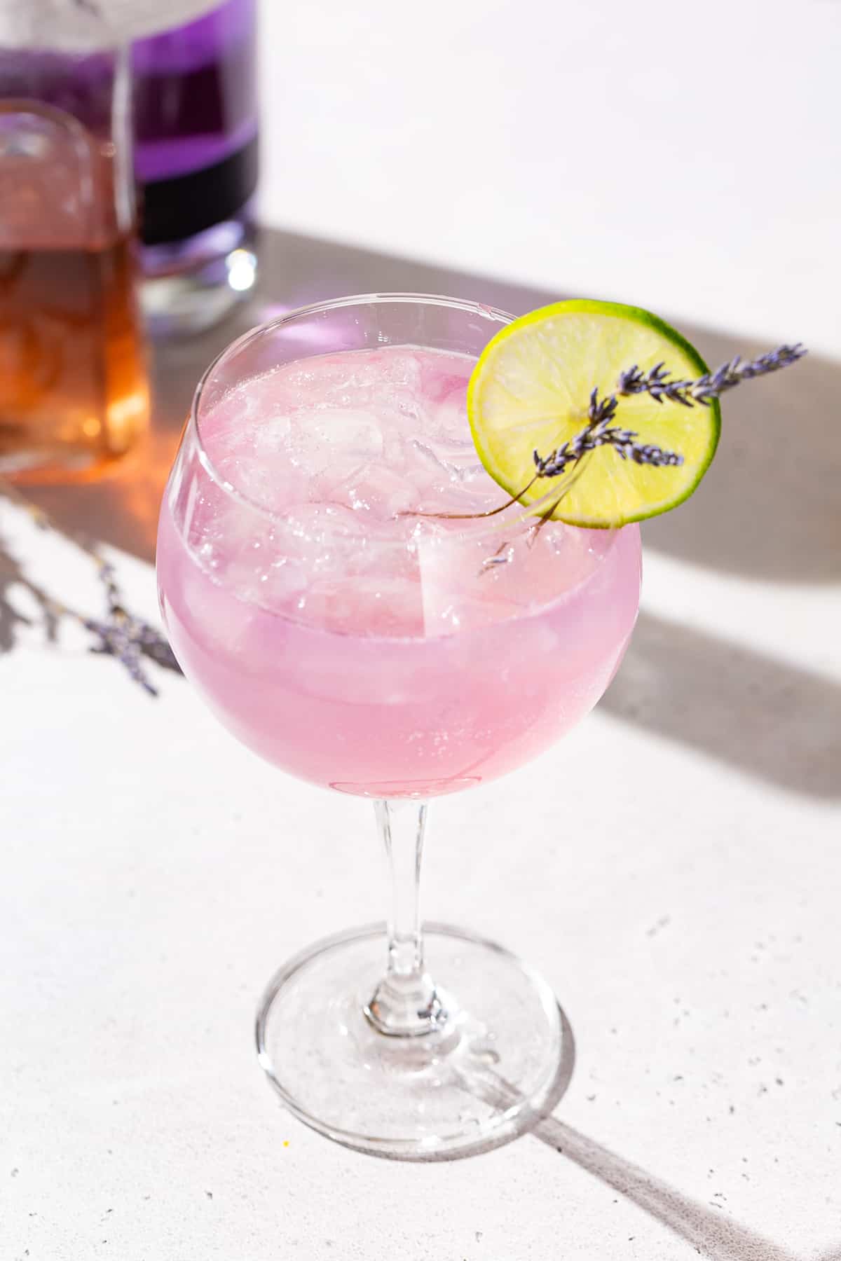 Lavender Lime gin spritz cocktail on a countertop with a bottle of lavender syrup and a bottle of purple colored Empress Gin in the background.