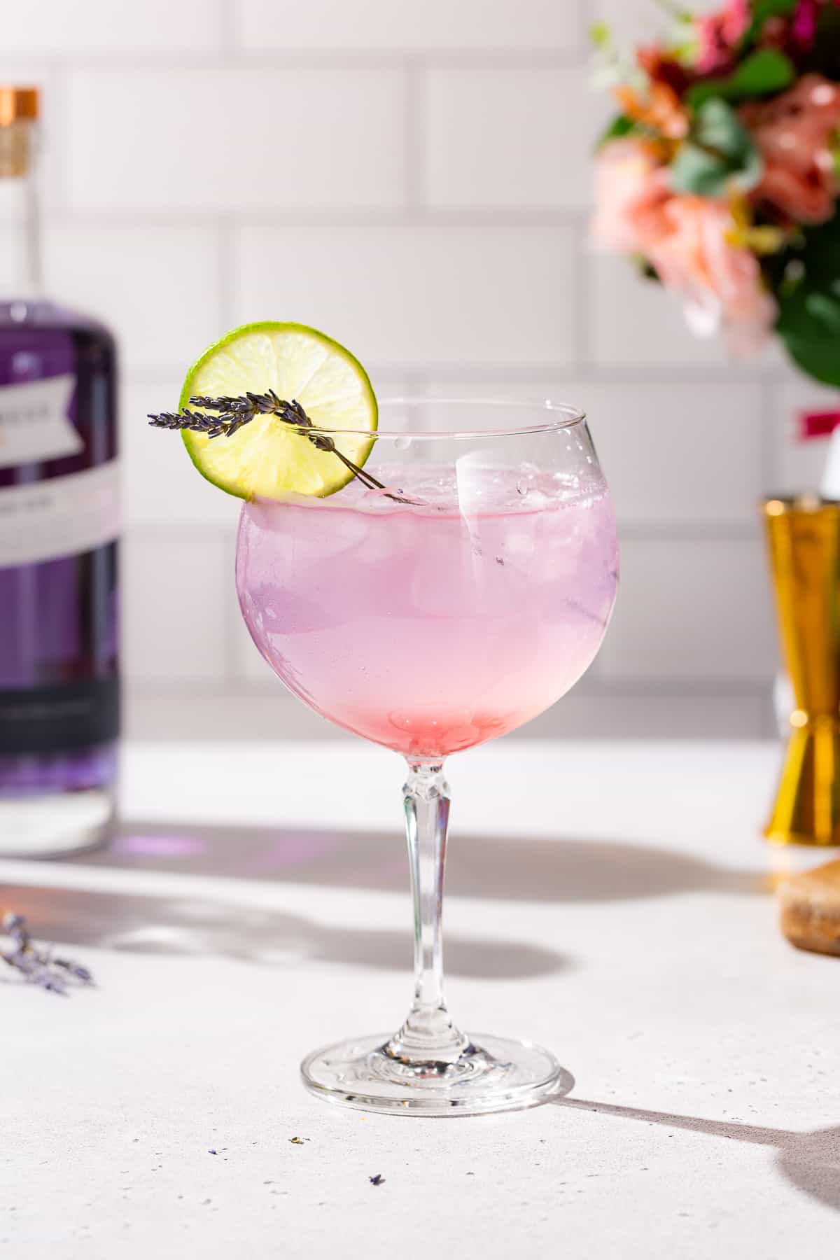 Side view of a Lavender Lime Gin Spritz cocktail. The drink is pinkish purple colored, with a brighter pink at the bottom of the glass. There is a lime slice and lavender sprigs on the rim of the glass as a garnish.