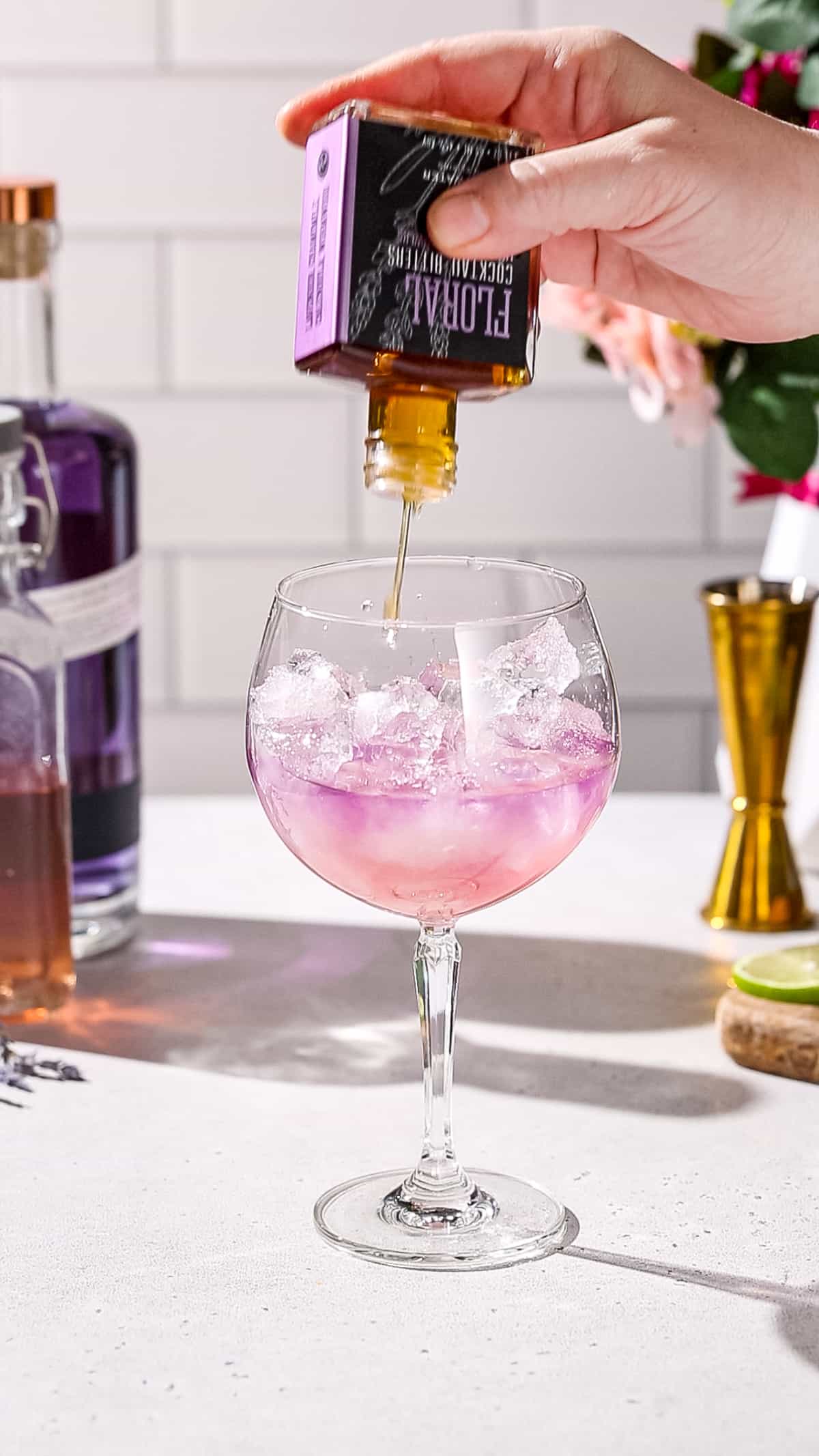 Hand adding lavender bitters to a cocktail glass filled with pink liquid.