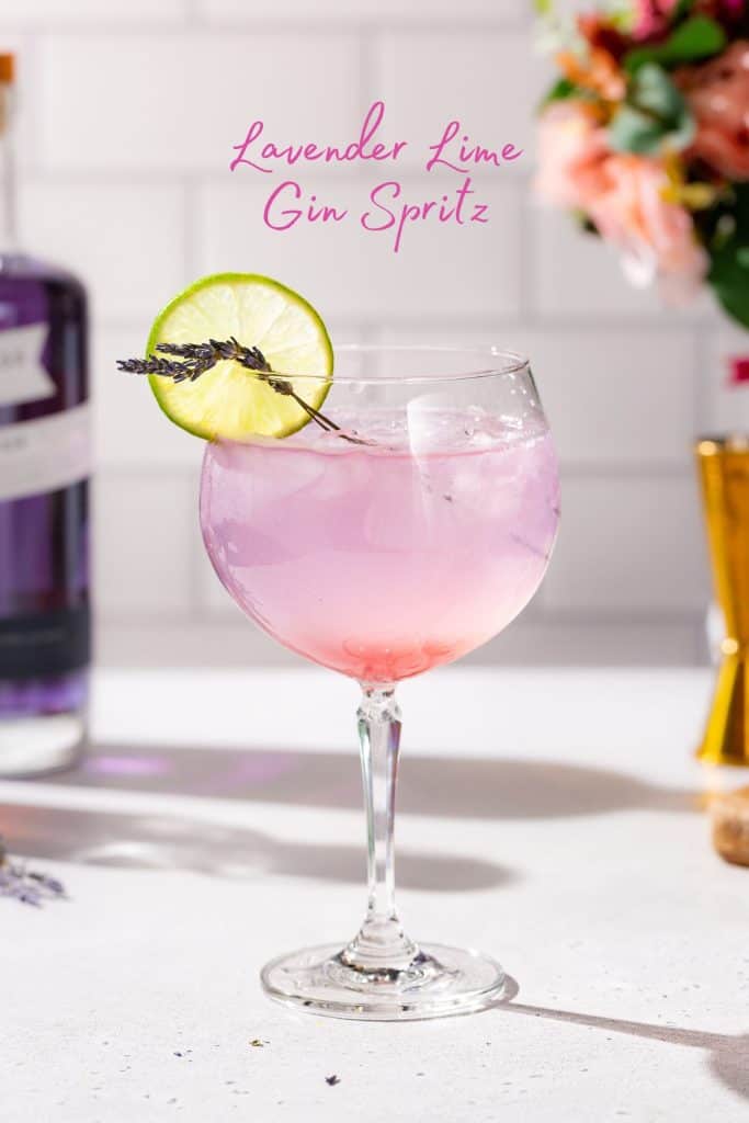 Lavender Lime gin spritz cocktail in a stemmed cocktail glass. The drink is a purple color and garnished with a lime slice and lavender sprigs. Text at the top says “Lavender Lime Gin Spritz”.