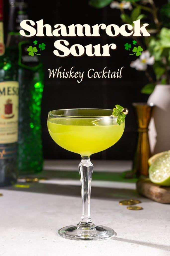 Side view of a green colored Shamrock Sour cocktail on a countertop. A bottle of Jameson, Midori and a lemon, lime and cocktail jigger are in the background. Text overlay at the top says “Shamrock Sour whiskey cocktail” along with some illustrated shamrocks.