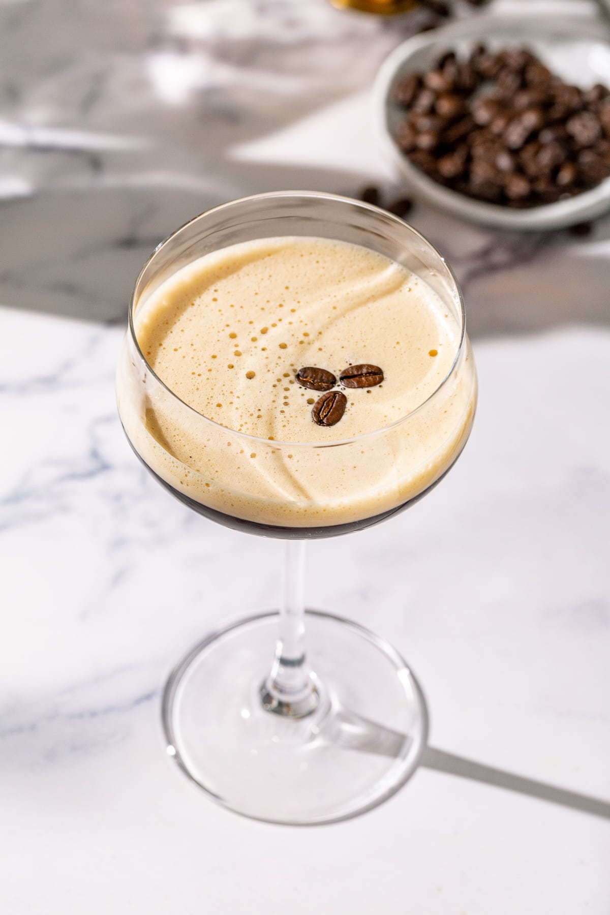 Overhead view of a Tequila Espresso Martini with three coffee beans as the garnish. in the background are more coffee beans. The drink is in a stemmed coupe glass and has a foamy layer on top.