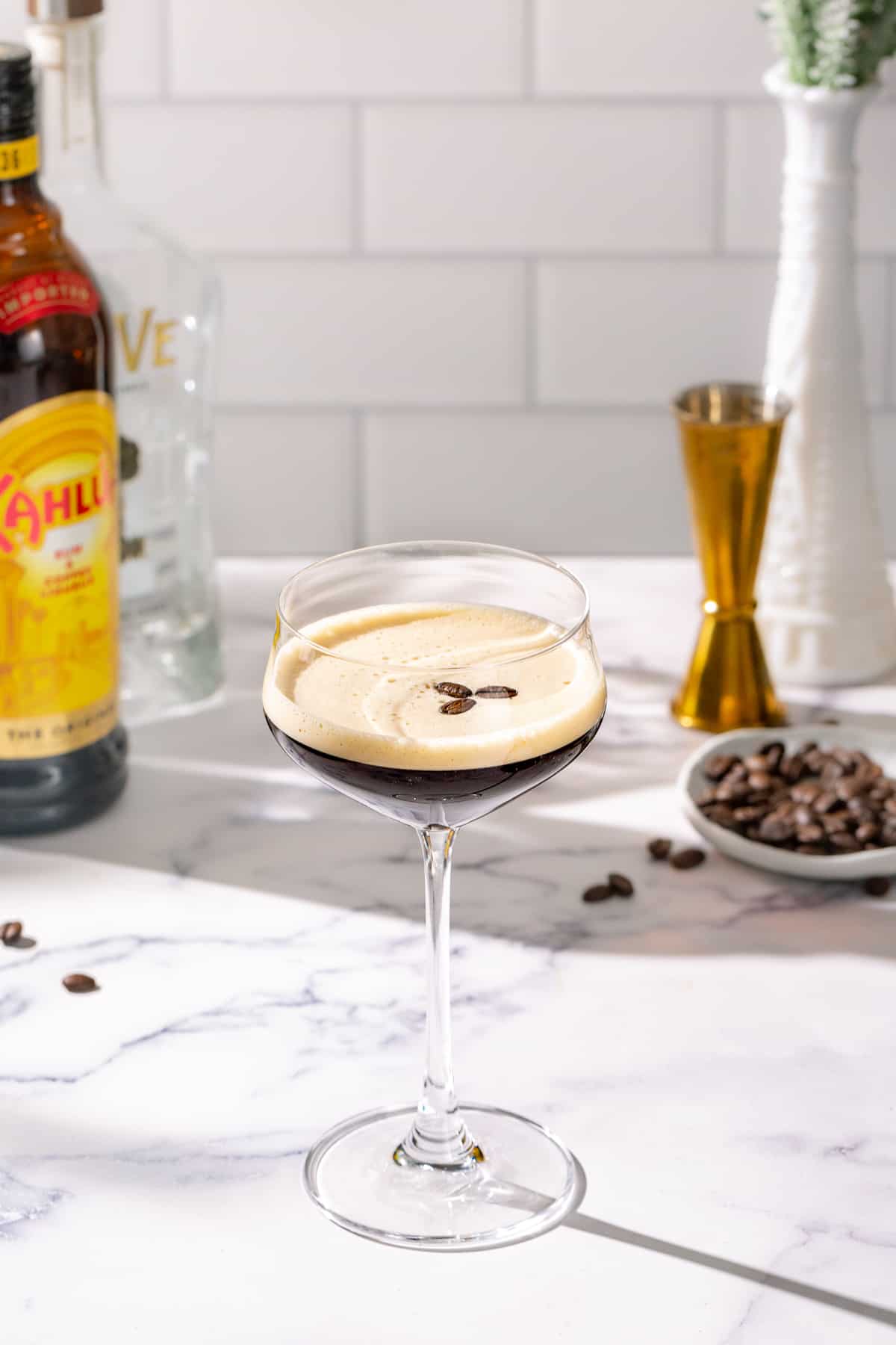 Tequila Espresso Martini on a marble countertop with a bottle of Kahlua coffee liqueur, a bottle of tequila blanco, coffee beans and a gold jigger in the background.