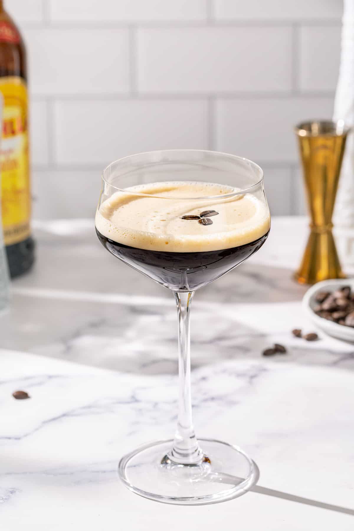 Tequila Espresso Martini on a marble countertop with a gold jigger, coffee beans and bottles of liquor in the background.