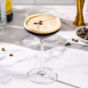 Tequila Espresso Martini on a marble countertop with a gold jigger, coffee beans and bottles of liquor in the background.