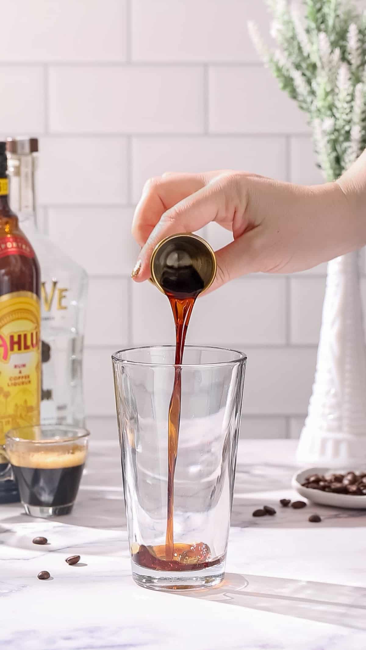 Hand pouring Kahlua coffee liqueur from a jigger into a cocktail shaker.