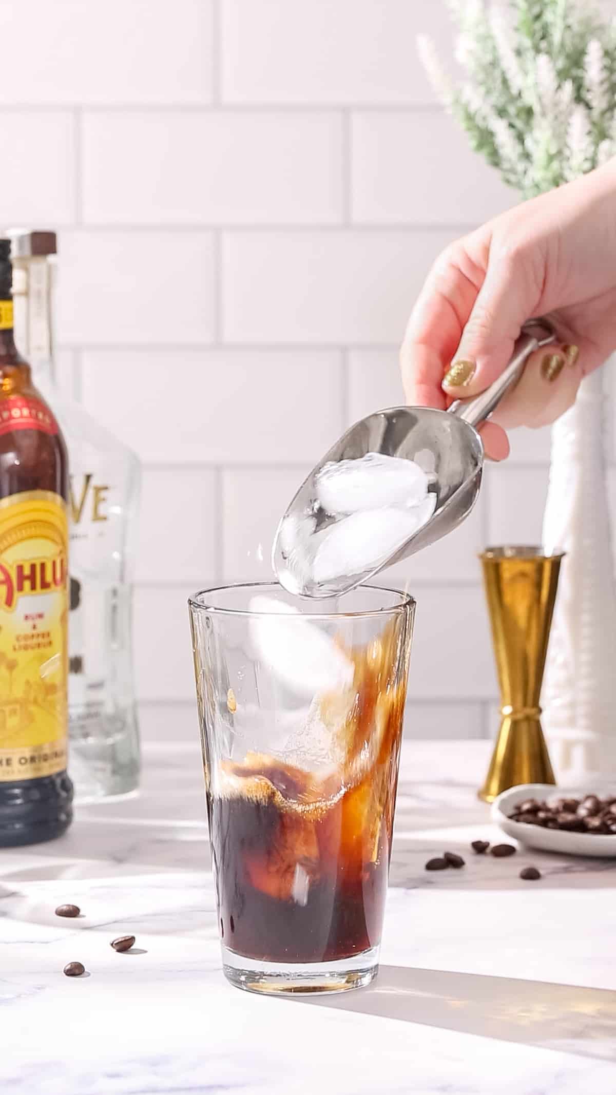 Hand filling a cocktail shaker with ice. The shaker also has dark brown liquid in it.