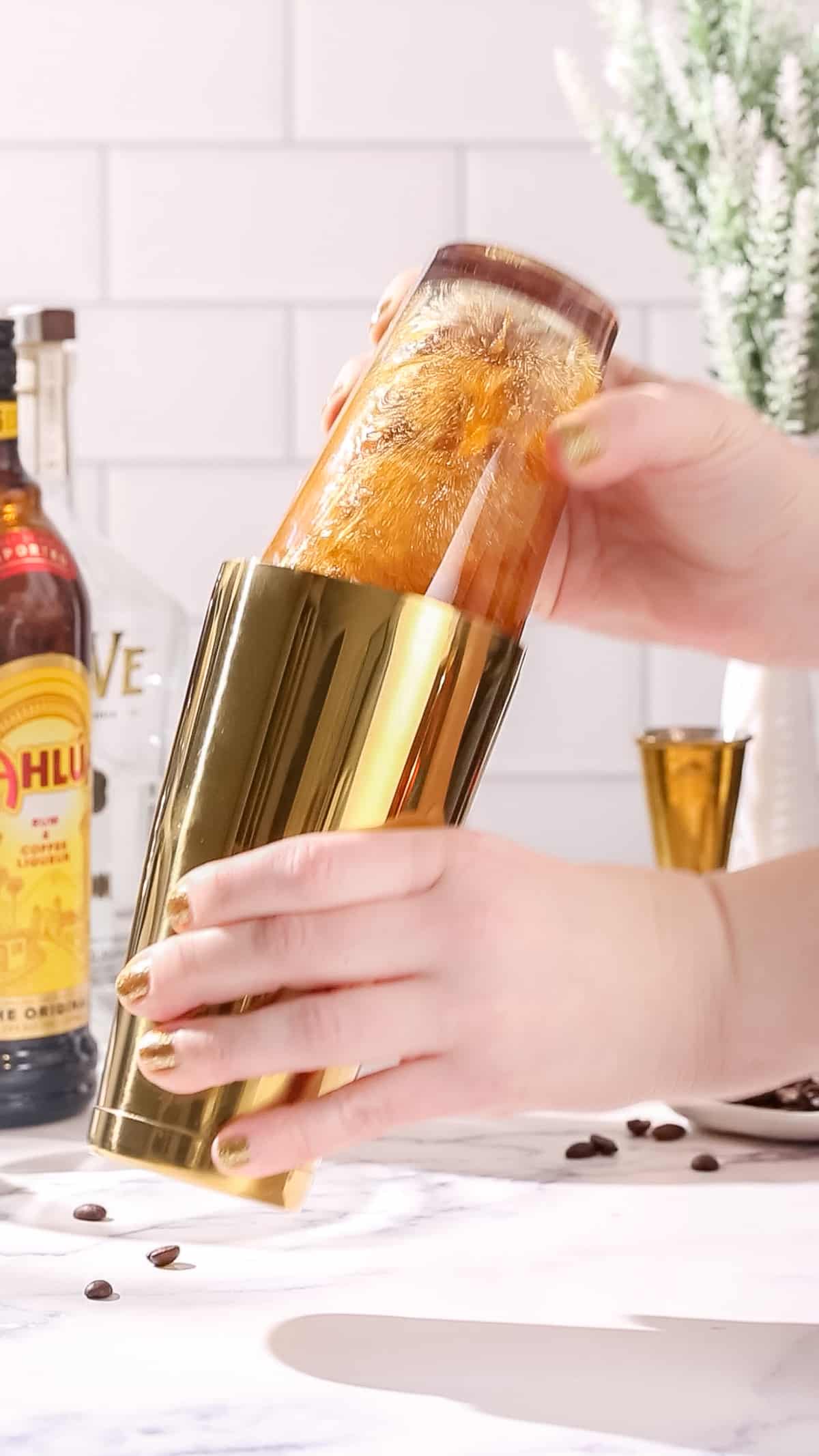 Hands shaking a gold and glass cocktail shaker filled with brown liquid.