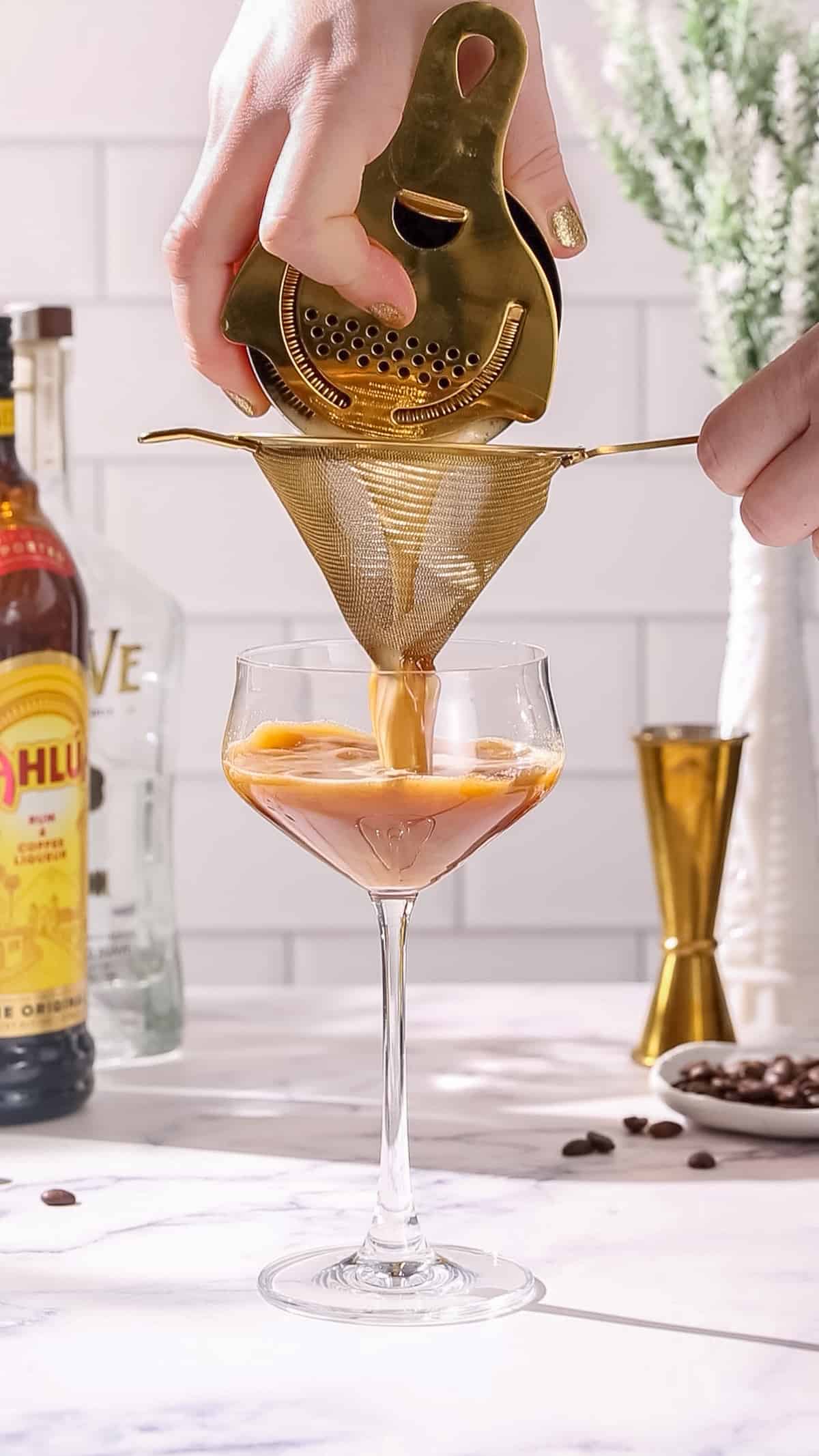 Hands straining a brown colored cocktail into a cocktail glass using a double straining method, where it is poured through a regular strainer at the same time as a fine mesh strainer.