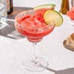 Frozen watermelon margarita on a countertop. The drink is garnished with a salt rim, a lime and a watermelon wedge. Bar tools and ingredients are in the background.