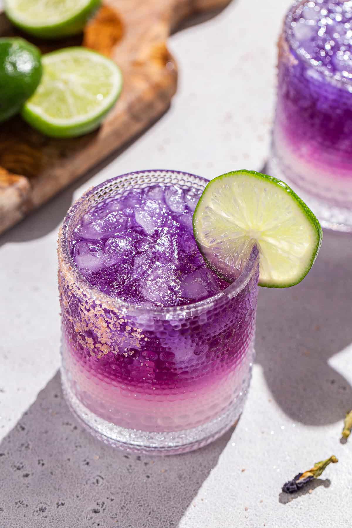 Overhead view of a purple layered margarita with a pink salt rim an a lime garnish. Another drink is in the background along with cut limes on a cutting board.