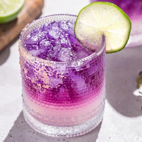 Close up of a Galaxy Margarita with another purple margarita and a lime in the background.