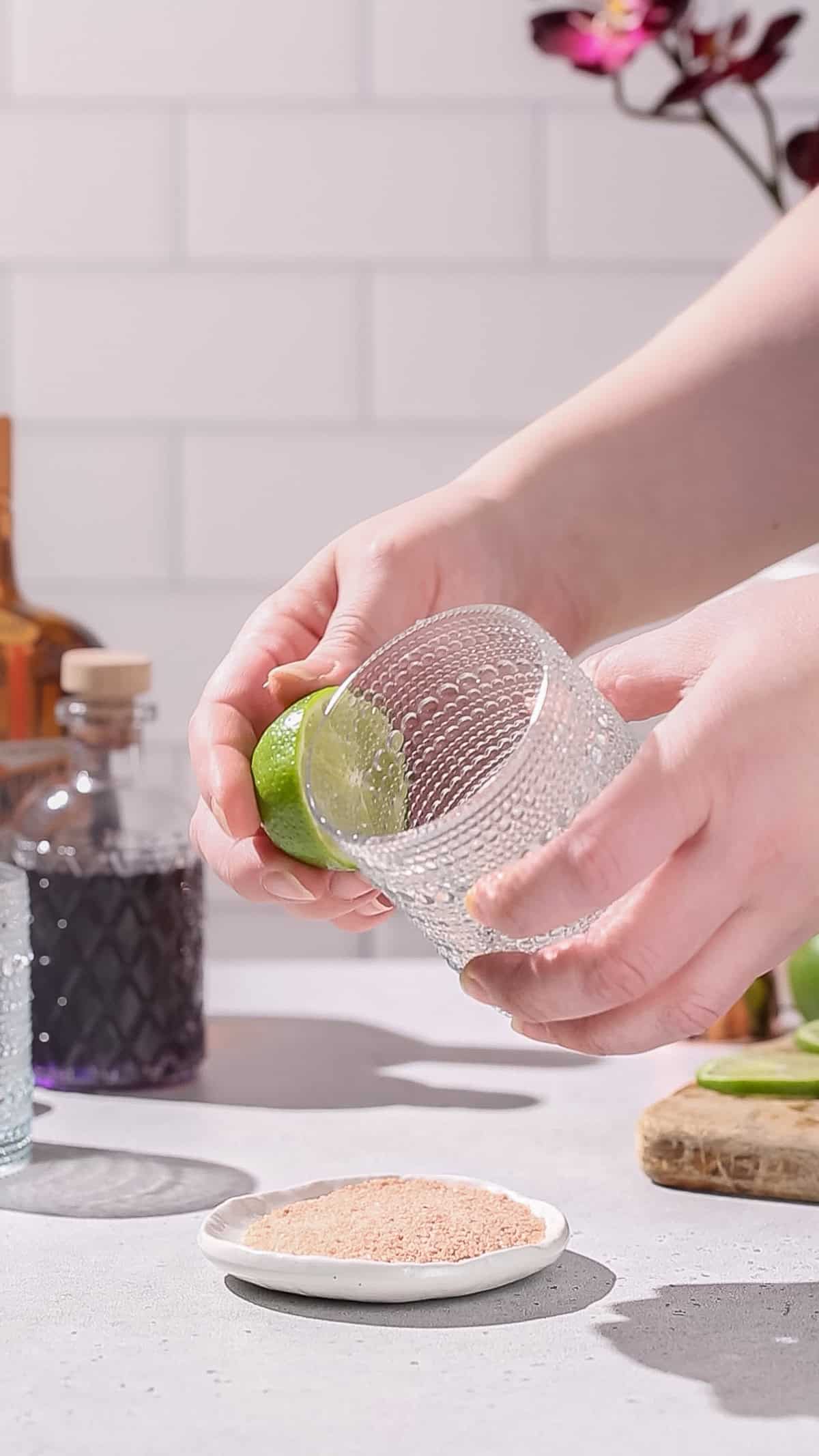 Hands using a cut lime to wet the rim of a cocktail glass.