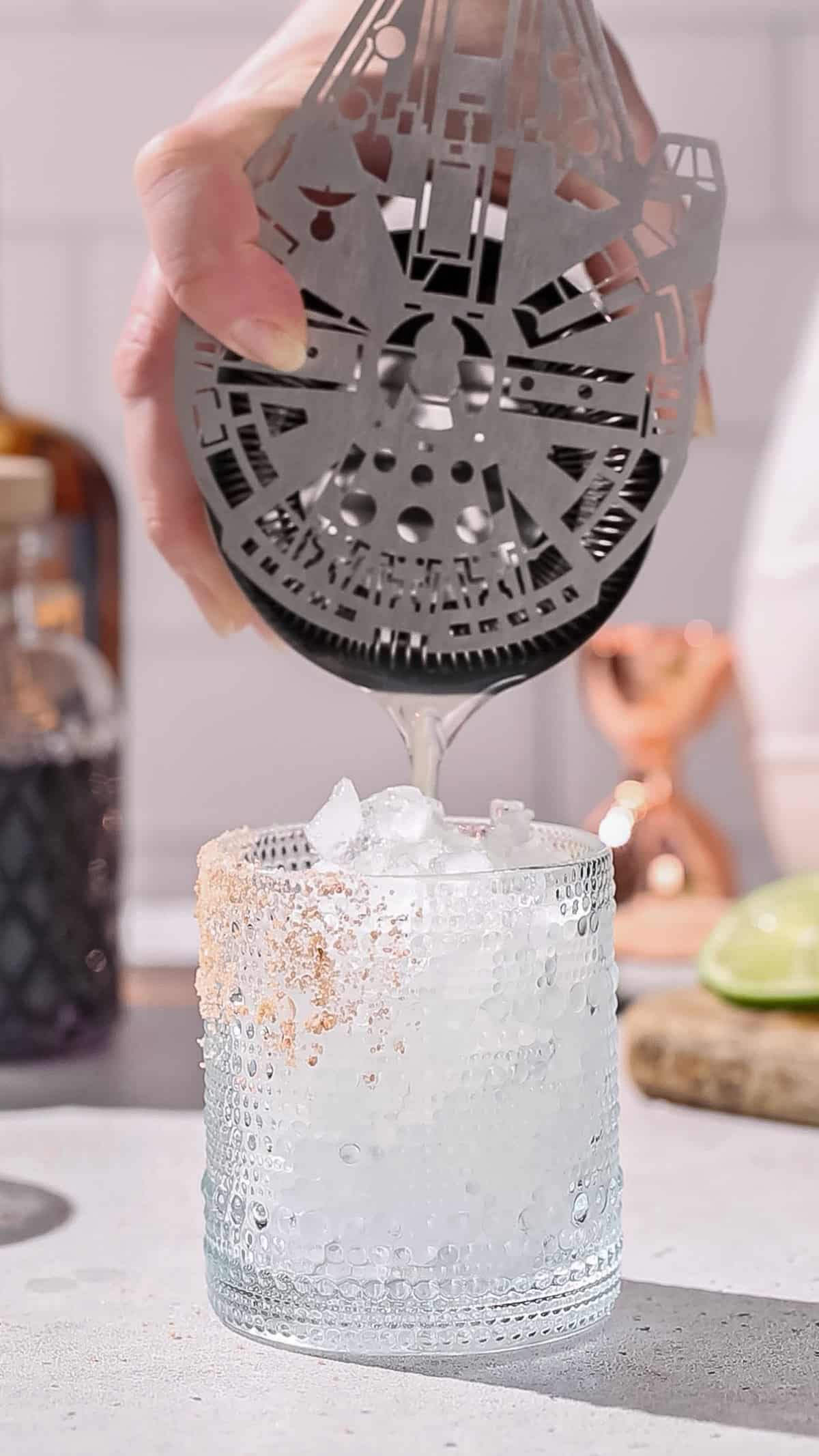 Hand using a Millenium Falcon cocktail strainer to pour liquid into a cocktail glass filled with ice.