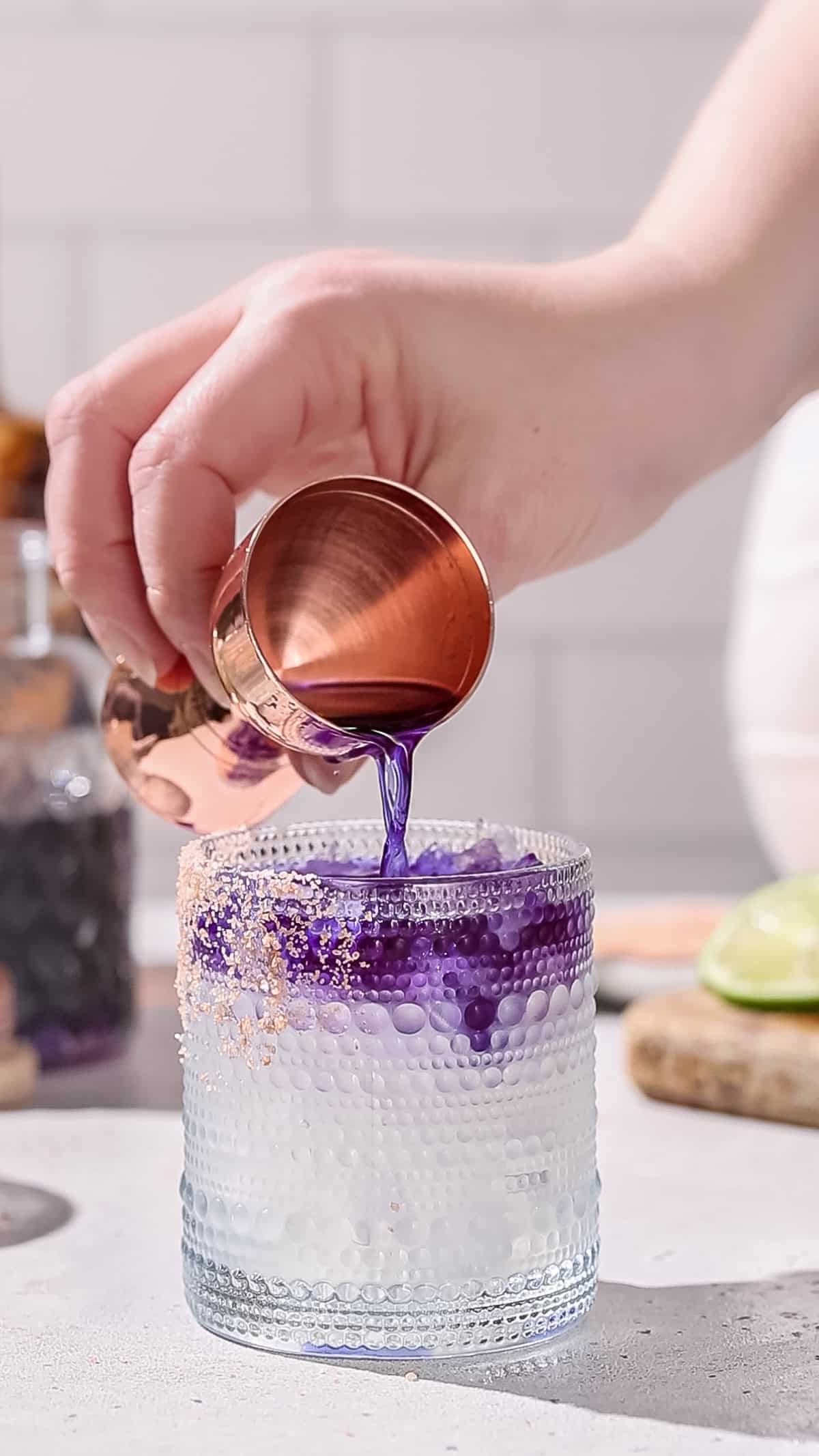 Hand using a jigger to pour purple liquid on top of a cocktail.