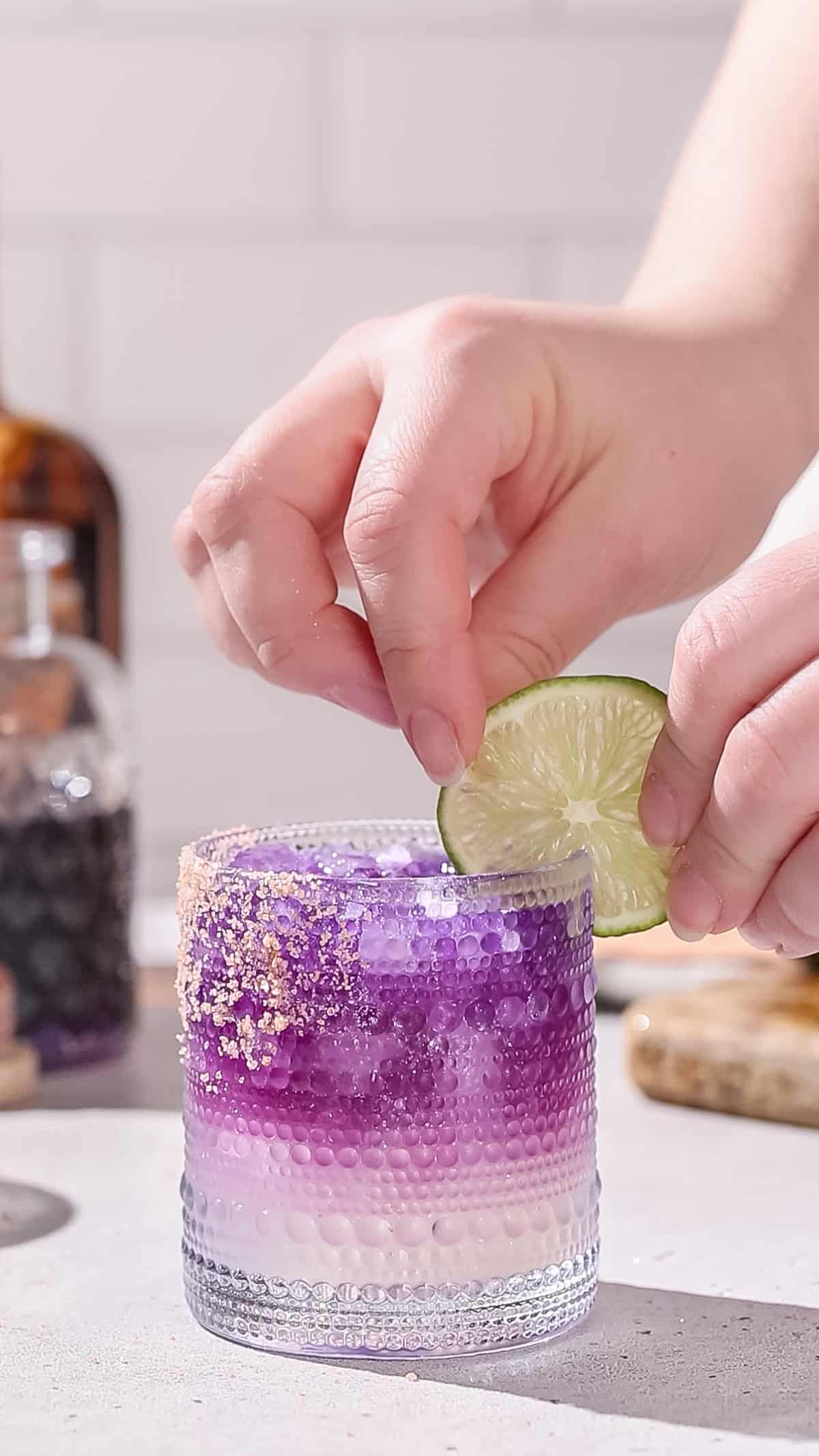 Hand adding a slice of lime to a purple cocktail.