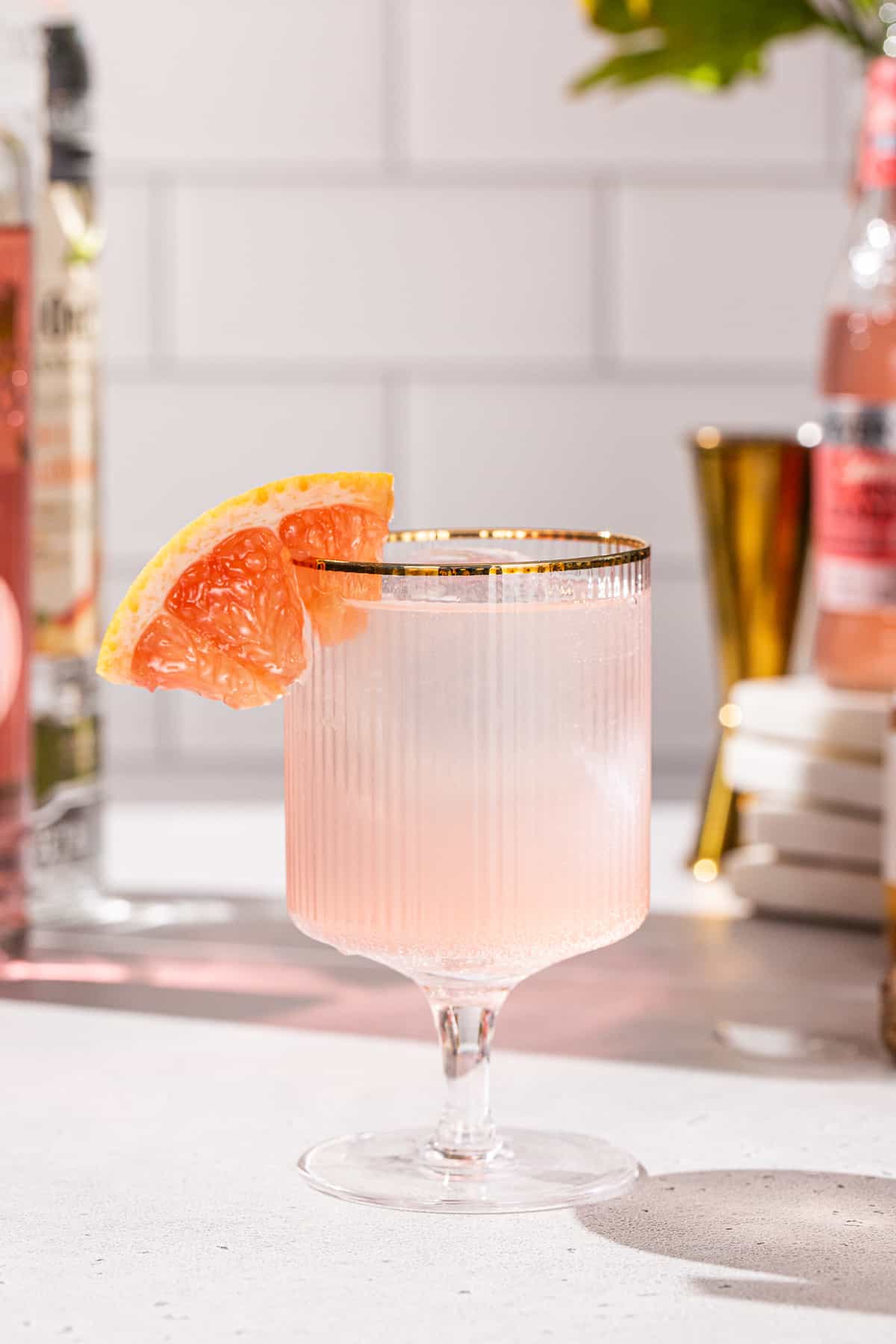 Side view of a Grapefruit Peach vodka cocktail, with a grapefruit wedge as a garnish, in a gold-rimmed cocktail glass. In the background are ingredients and bar tools to make the drink.