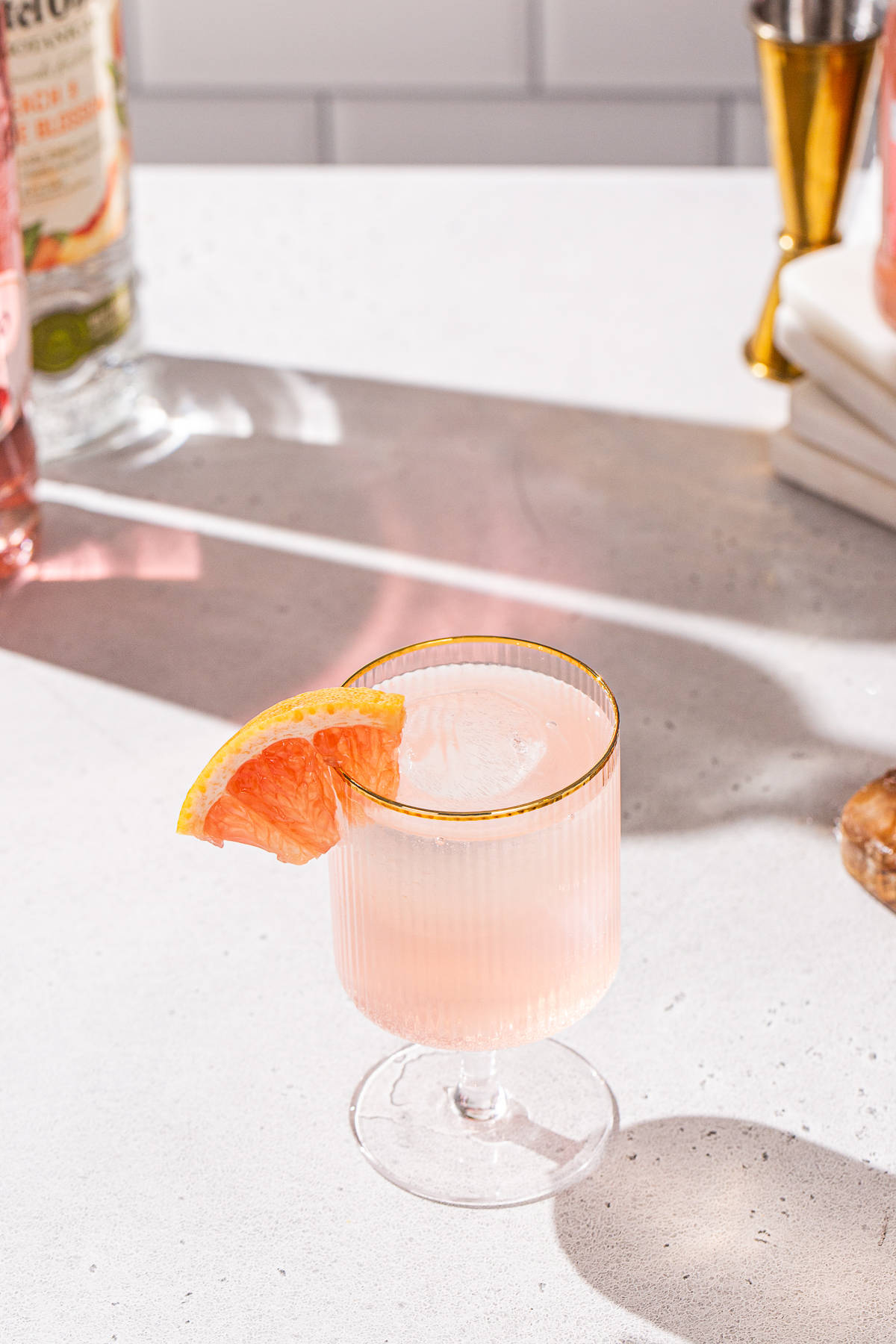 Overhead view of a gold-rimmed cocktail glass with a peach colored cocktail that has a piece of fresh grapefruit as a garnish. In the background are bottles used in the drink.