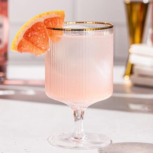 Close up of a gold-rimmed ribbed cocktail glass with a short stem that contains a peach colored cocktail. The drink is garnished with a piece of fresh grapefruit.