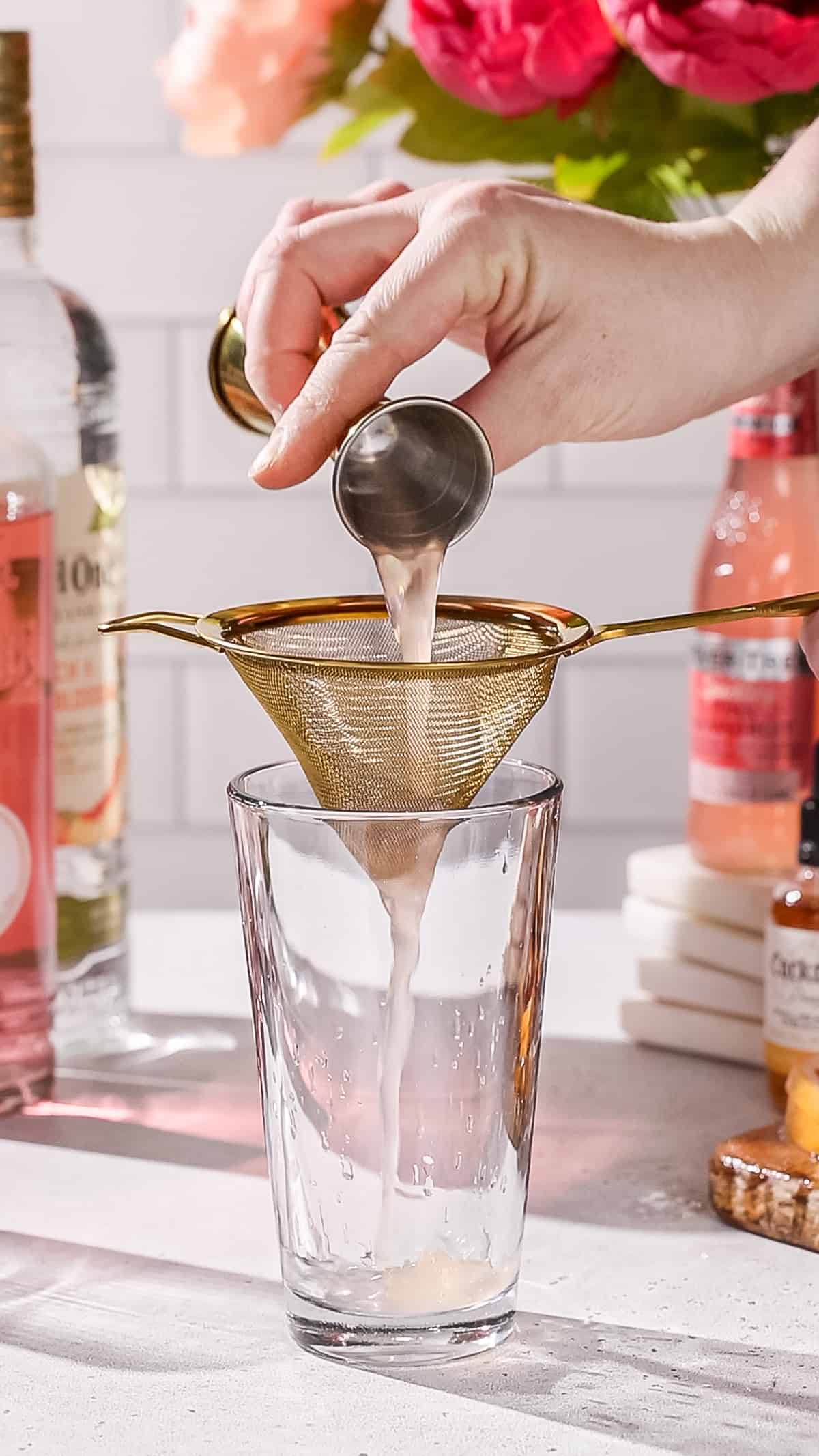 Hand using a fine mesh strainer to pour grapefruit juice into a cocktail shaker.