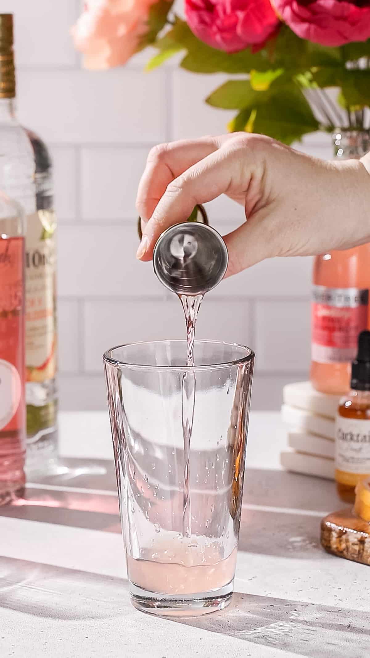 Hand pouring pink lychee liqueur into a cocktail shaker.