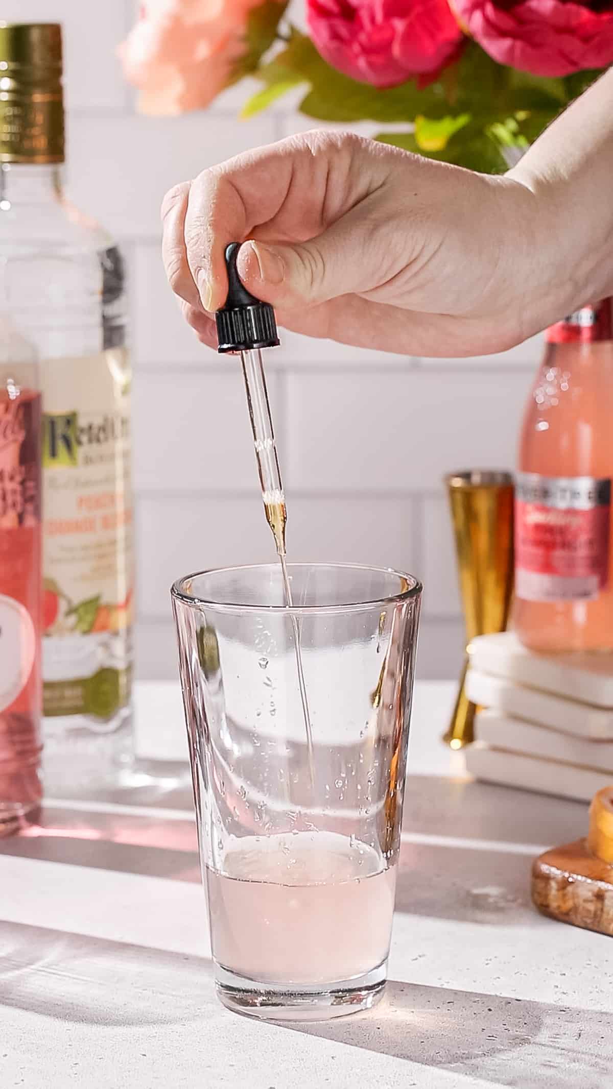 Hand using a dropper tool to add peach bitters to a cocktail shaker.