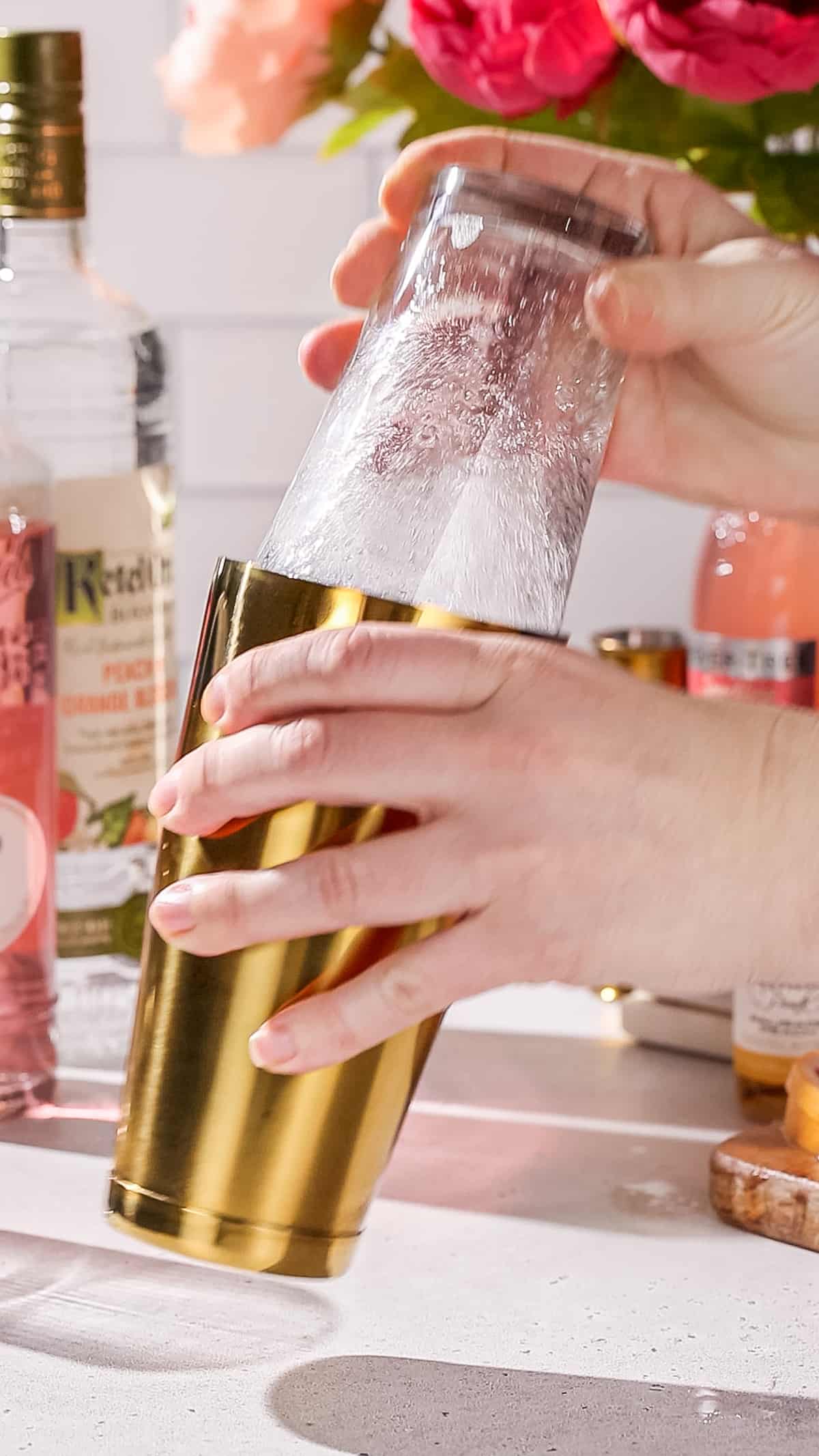 Hands shaking a cocktail using a glass and gold cocktail shaker.