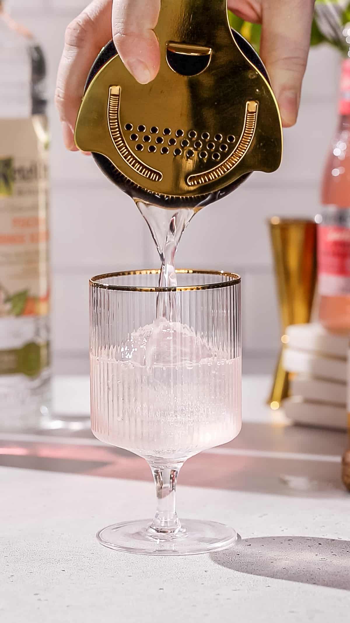 Hand straining a cocktail into a gold-rimmed glass.