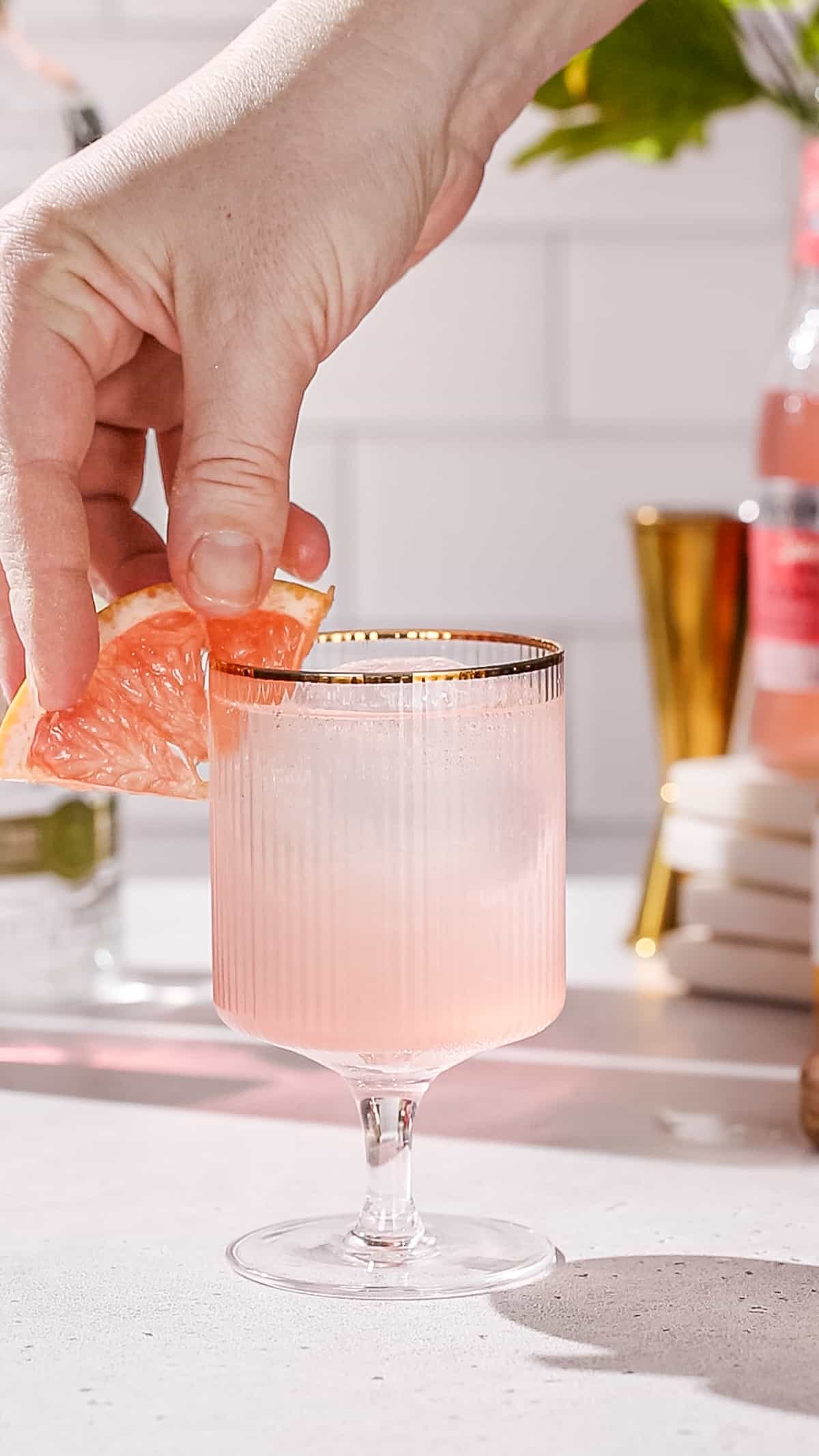 Hand adding a piece of pink grapefruit to a cocktail glass as a garnish.