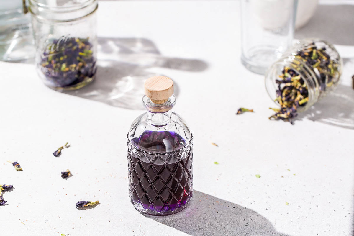 Bottle of purple butterfly pea tequila on a countertop with two jars of butterfly pea flowers in the background.