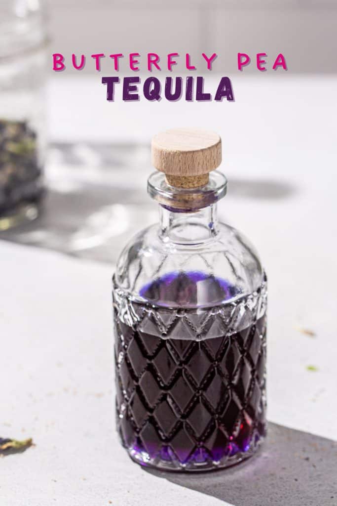 Overhead view of a bottle of purple butterfly pea tequila on a countertop with butterfly pea flowers in the background. Text overlay says “Butterfly Pea Tequila”.